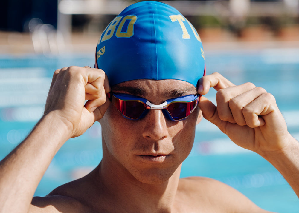 Person wearing a blue swim cap and red goggles by a pool