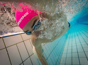 Person swimming underwater with a pink swim cap and goggles in a pool.