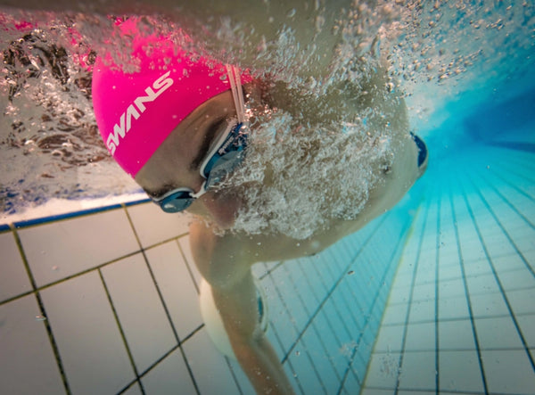 Person swimming underwater with a pink swim cap and goggles in a pool.